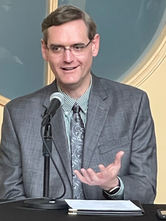 A man with grey hair and glasses speaking into a microphone. He wears a light blue-grey suit and tie.