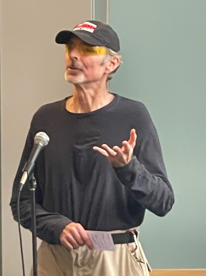 An older man with grey hair and a black ball cap wearing a black shirt stands at a microphone and speaks into it.