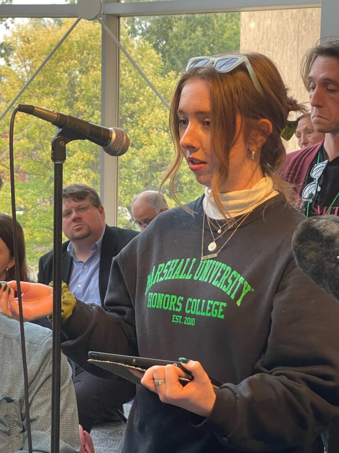 A young woman, college age, with brown hair and sunglasses, wearing a black Marshall University sweatshirt stands a microphone and speaks into it.