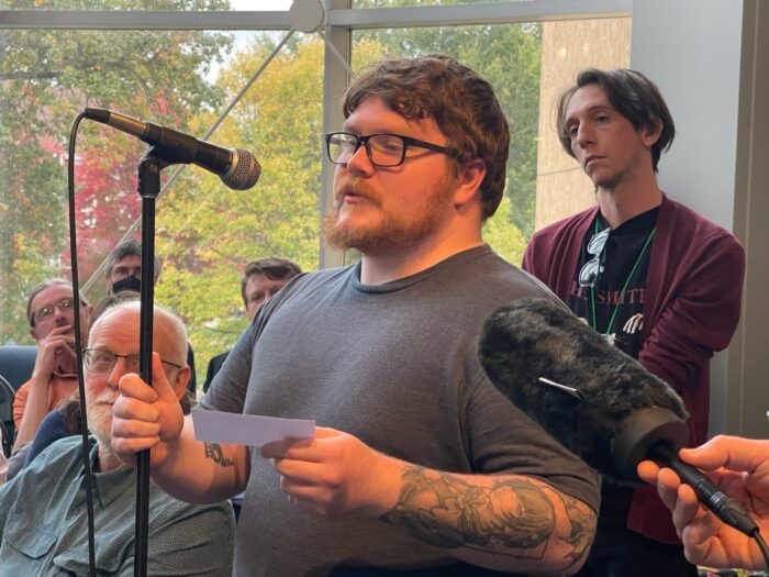 A millennial age man with red-brown hair, wearing glasses and with tattoos on his forearm, stands at a microphone and speaks into it.