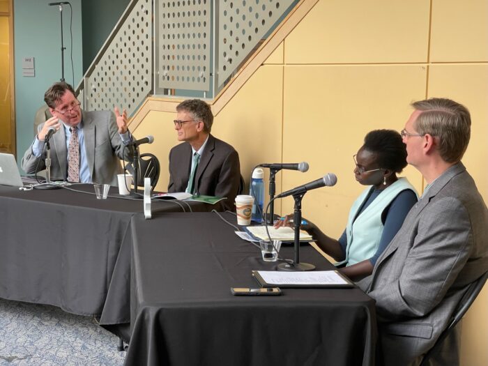 Four people sit at two tables with black tablecloths for a panel discussion. Each person has a microphone.
