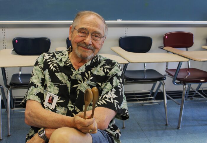 An older man sits in a chair in a classroom while holding wooden spoons.