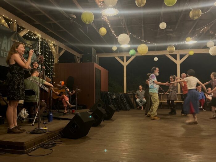 A woman speaks into a microphone on a stage. People square dance on the dance floor.