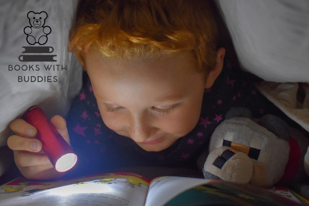 A child with red hair reads a children's book under a blanket while holding a stuff animal. The child is smiling and holding a small, pink flashlight. In the corner of the image is a logo with the words "Books with Buddies." Above the text is a little teddy bear sitting on a stack of books.