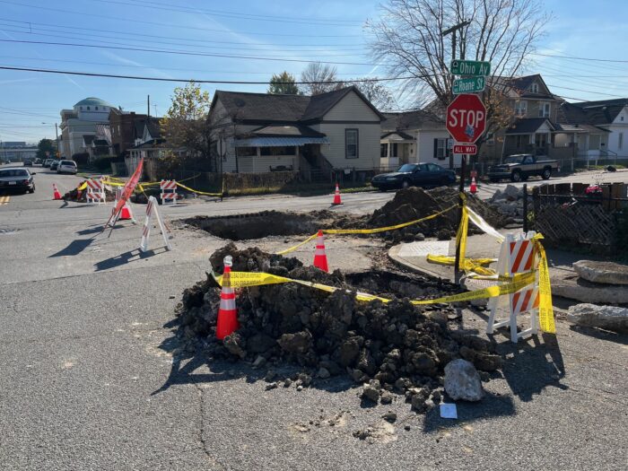 Holes in the street are surrounded by mounds of dirt, yellow tape and orange cones.
