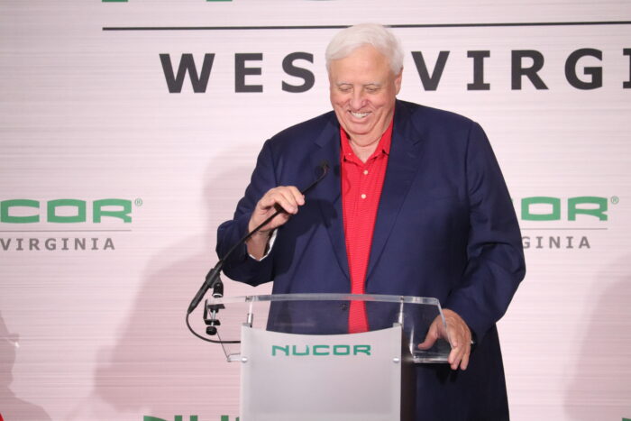 A big man with white hair, wearing a blue jacket and red shirt, speaks to a crowd of people under a tent.