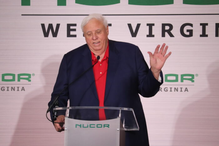 A large man with white hair wearing a blue jacket over a red shirt speaks to an audience from a stage under a tent.