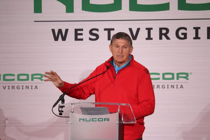 A man wearing a red jacket speaks to an audience from a stage under a tent.