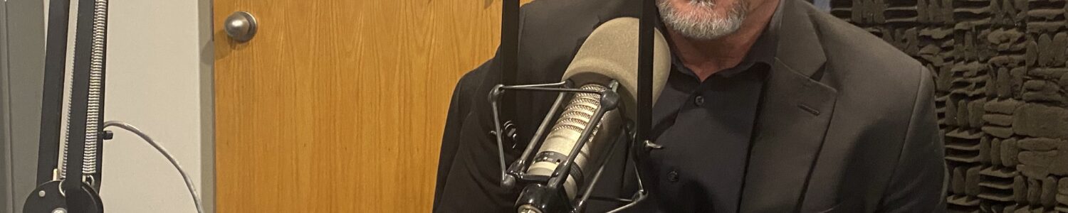 Bald man with beard dressed in black at studio microphone.