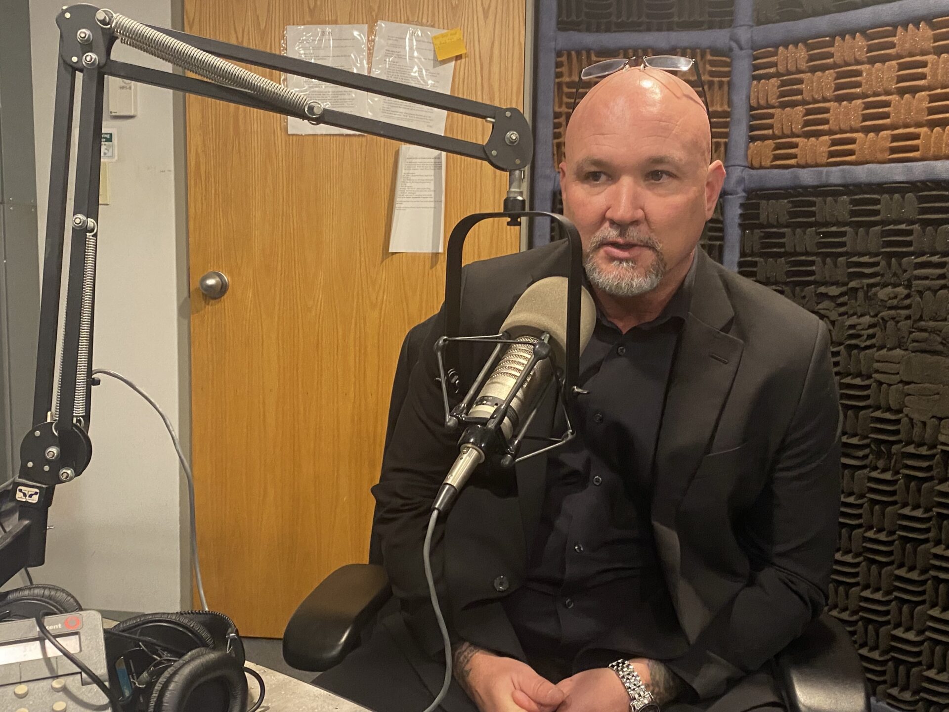 Bald man with beard dressed in black at studio microphone.