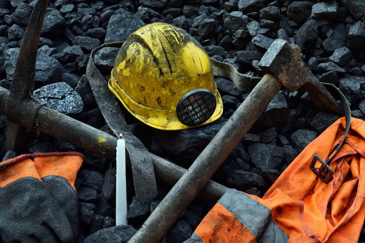 A yellow miner's hat is placed on top of pieces of coal, with mining tools and an orange safety jacket.