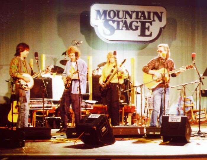 Four people stand on a stage singing and playing instruments. The Mountain Stage logo is seen behind them on a wall. 
