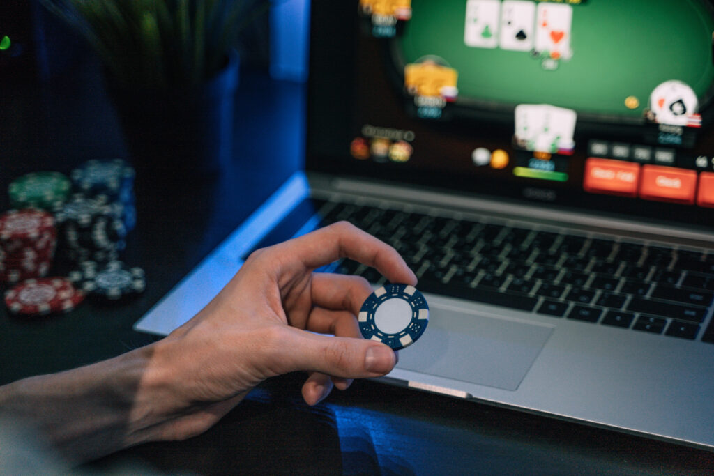 A hand holding a poker chip in front of a computer screen showing online poker being played