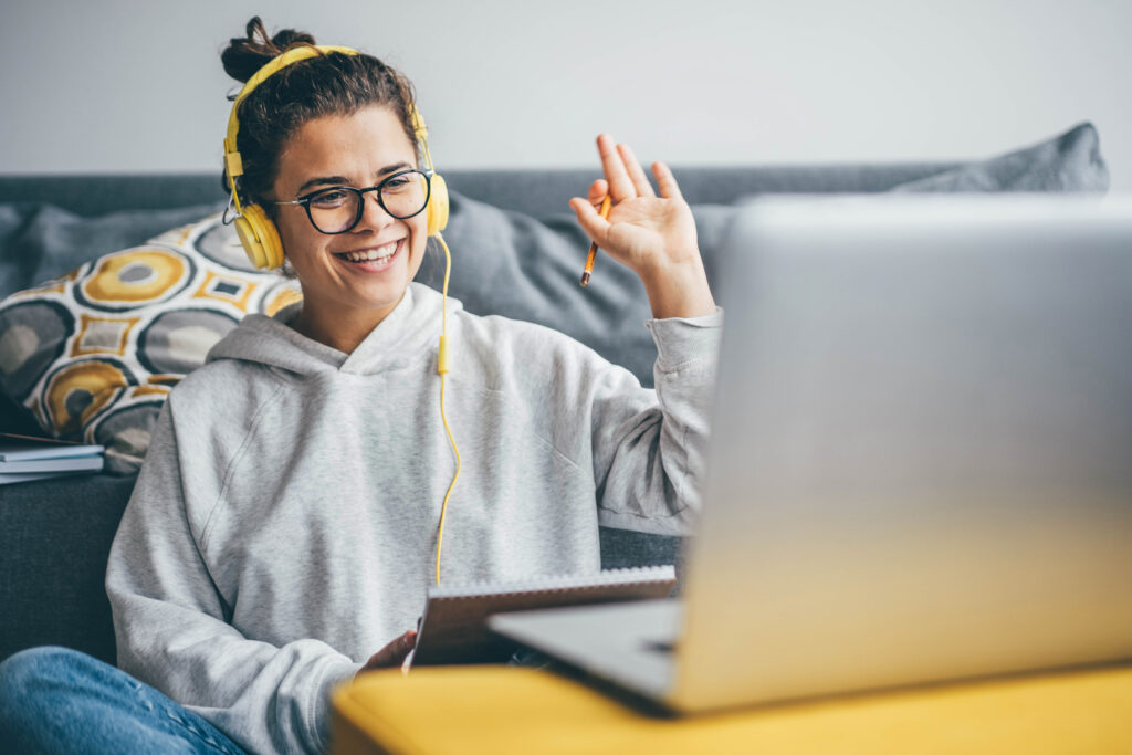 A millennial age woman waves her hand saying hello on a video call on her computer at home. The woman had yellow headphones and wears a gray hoodie and black glasses.