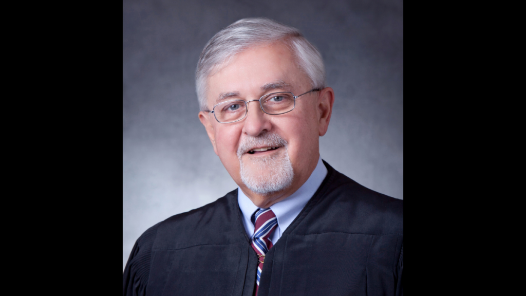 White haired man with glasses and goatee in Judge's robes.