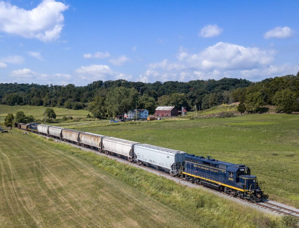 A train pulled by a blue locomotive pulls through a green rural setting under a blue sky with white clouds.