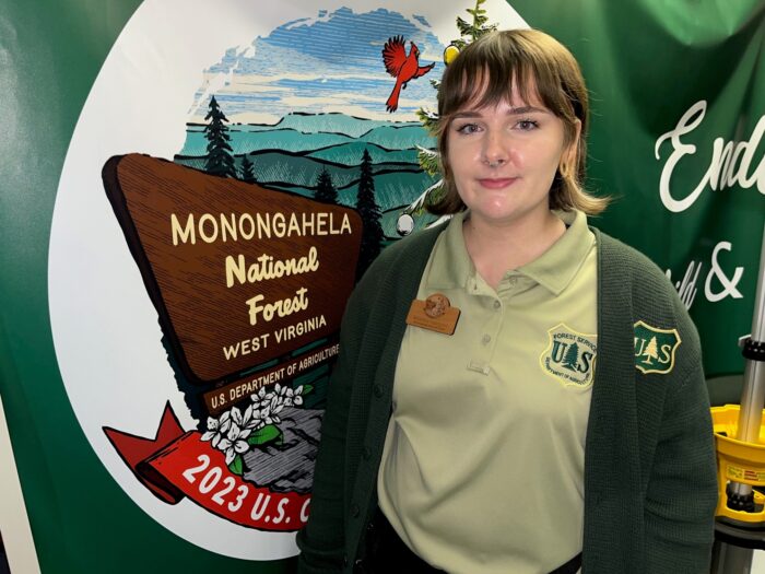 A young woman wearing a green jacket stands under a tent next to a picture of a sign for the Monongahela National Forest.