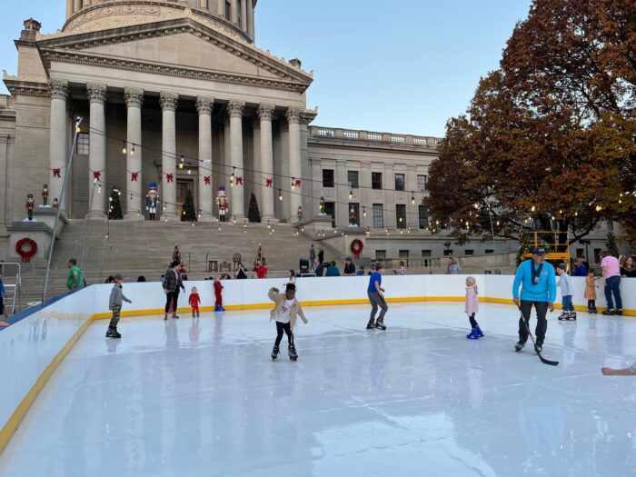 Children and adults skate in a rink in front of a stone building with large columns.