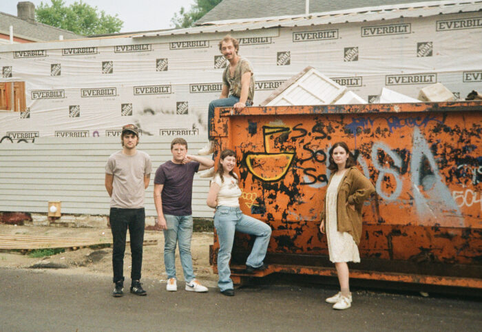 Four people stand in front of an orange dumpster.
