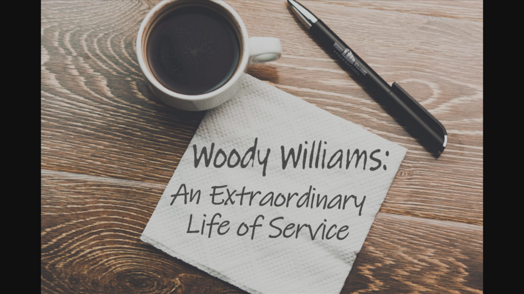 A coffee cup from a above is shown on a table. A napkin with a pen next to it reads, "Woody Williams" An Extraordinary Life of Serivice."