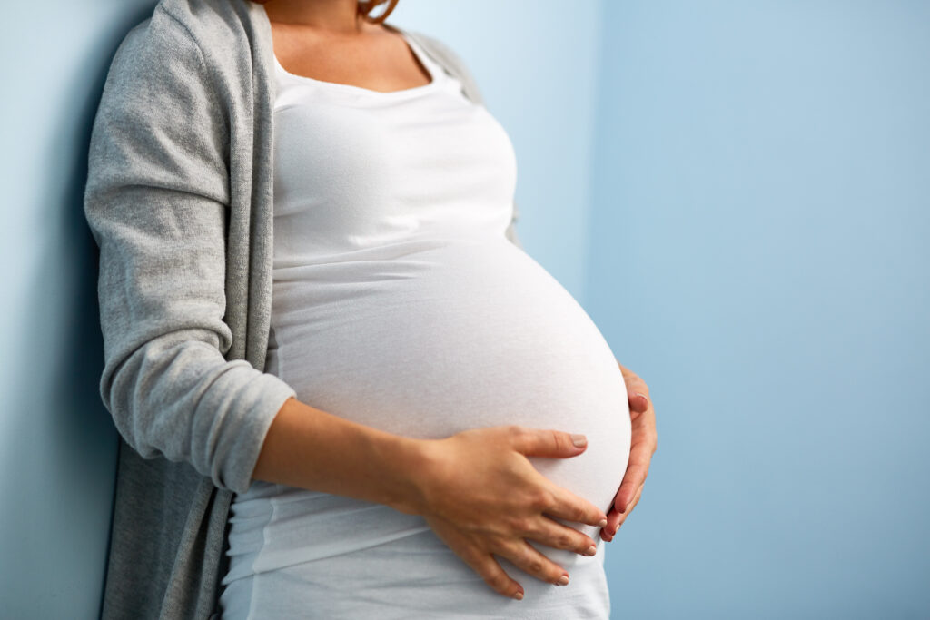 A pregnant woman holds her belly. She wears a white shirt and a gray cardigan.