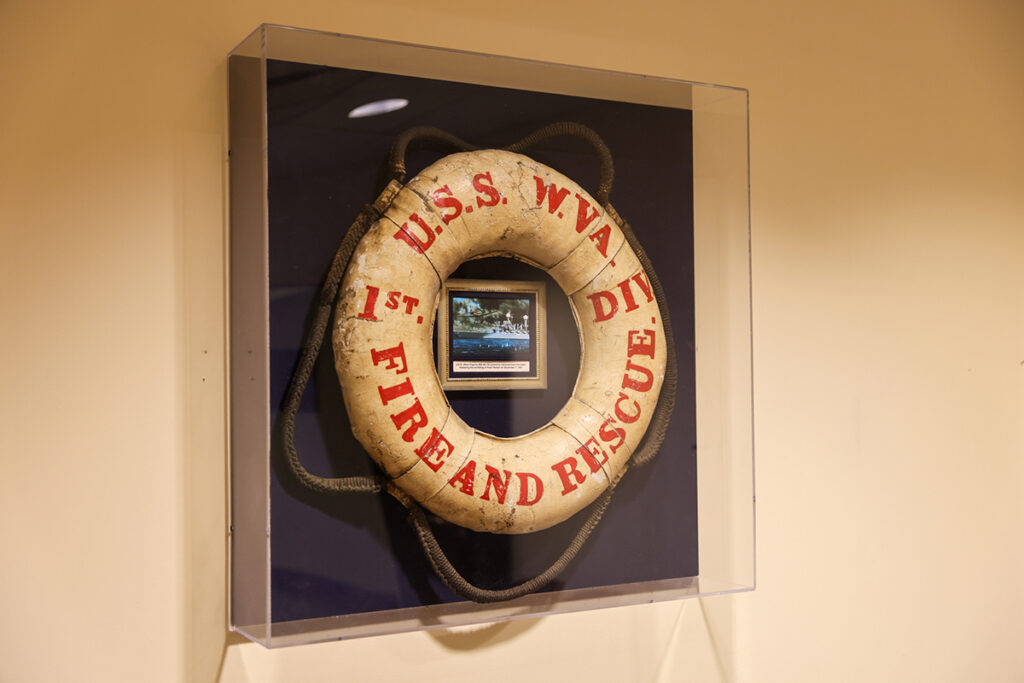 A glass display case mounted to a white wall shows a white life preserver with a decorative rope on a navy blue backing. Red writing on the life preserver reads "U.S.S. W.VA. 1st DIV. FIRE AND RESCUE". Inside the life preserver can be seen an image of the USS West Virginia.