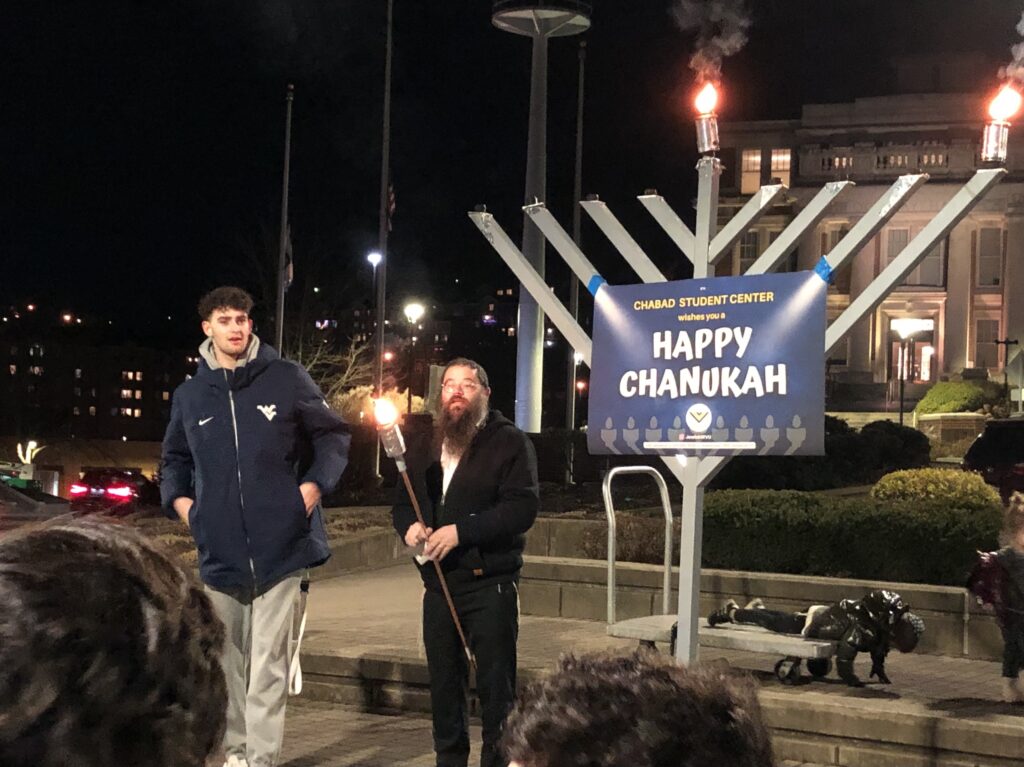Two men stand next to a large menorah, one of them holding a torch. The menorah has two of its nine branches lit, and has a sign hung from it that reads "Happy Chanukah"