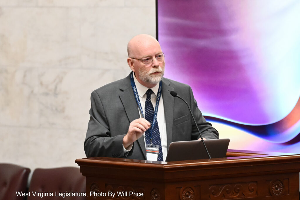 A man dressed in a grey suit over a white shirt and dark blue tie stands at a lectern in the West Virginia Senate chambers. He is gesturing with his left hand as he speaks into a slender microphone. He stands in front of a large purple screen, which is in turn in front of a white marble wall.