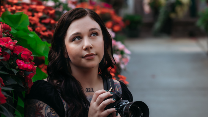 An adult woman with dark hair and tattoos poses for a photo and looks up toward the sky, while holding her camera. A flower bush is seen behind her.