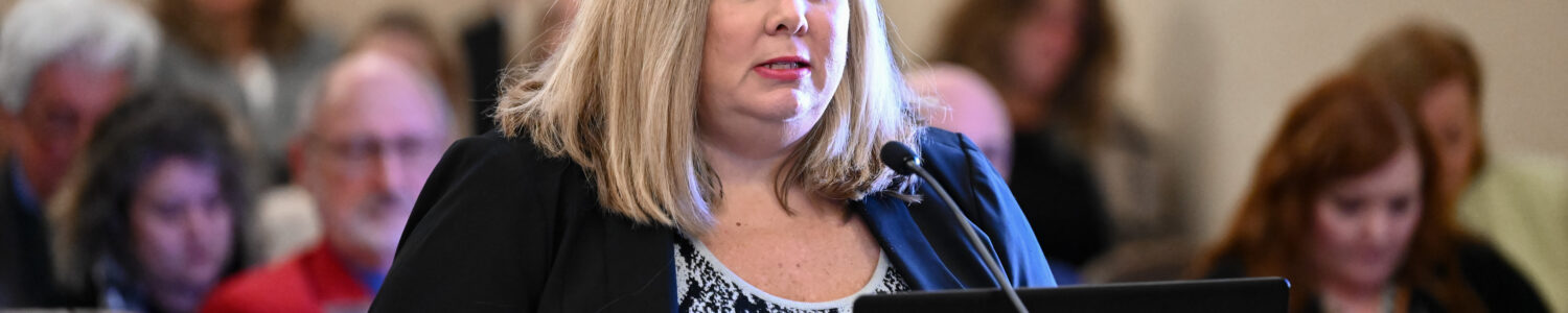 A blonde woman wearing a black blazer speaks from a podium.