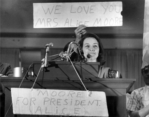 A black and white photo of an adult woman. She stands at a podium holding a sign up that reads, "We love you Mrs. Alice Moore."