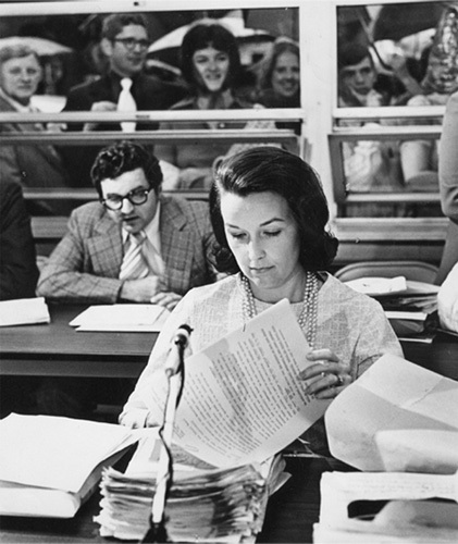 A black and white photo of an adult woman sits at a desk. A microphone is in front of her along with a large stack of papers.