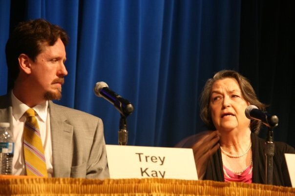 Two adults are shown in a photograph. On the left is a man with a short beard and a mustache. On the right is an older woman. Both appear to be speaking on a panel.
