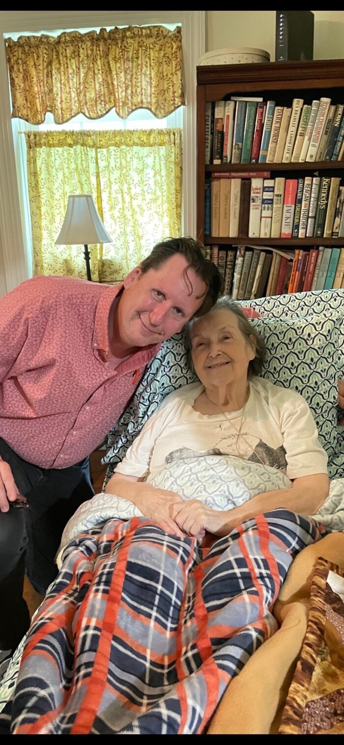 A photograph of an adult man and an elderly woman. The man leans down toward the woman who sits in a chair covered in blankets.