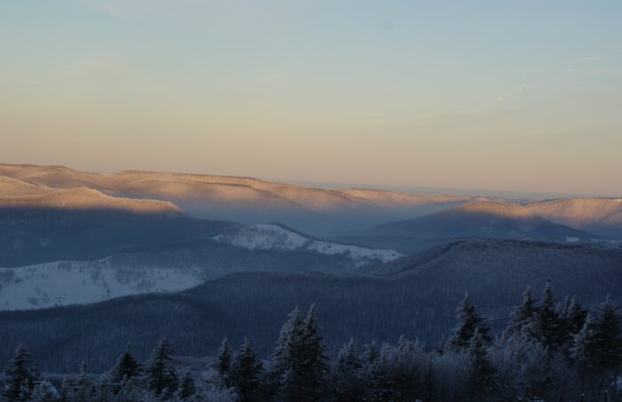 A shadow covers much of the image with frosted evergreens in the foreground and snow-dappled hills rolling away into a winter landscape where a faint, pink sunlight touches the distant peaks.