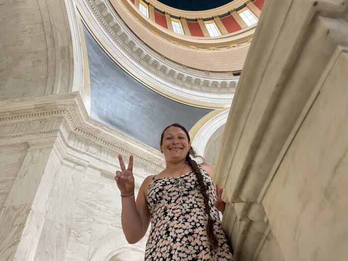 A middle age woman wearing a floral dress stands in the WV State Capitol Rotunda making a peace sign with her right hand.