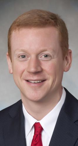An adult man with red, short hair, wearing a suit and tie smiles for a photo.