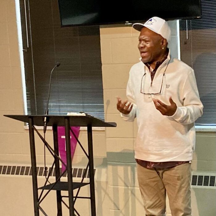 A middle age Black man wearing a white pullover jacket and ball cap speaks from a podium.