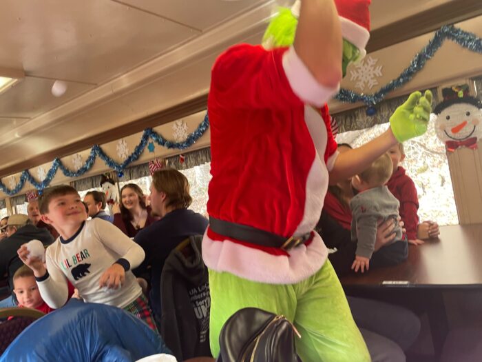 A man dressed as the grinch moves through a crowded train as a kid throws a fake snowball at him. 