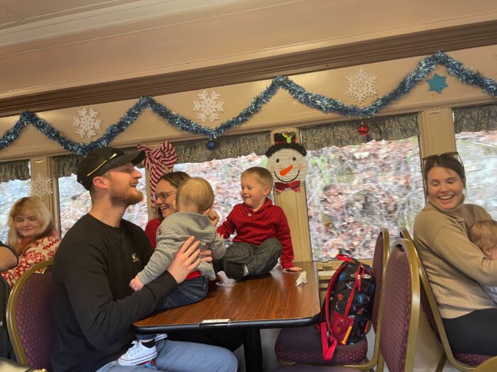 Families sit on a table on the train holding babies and smiling at each other.