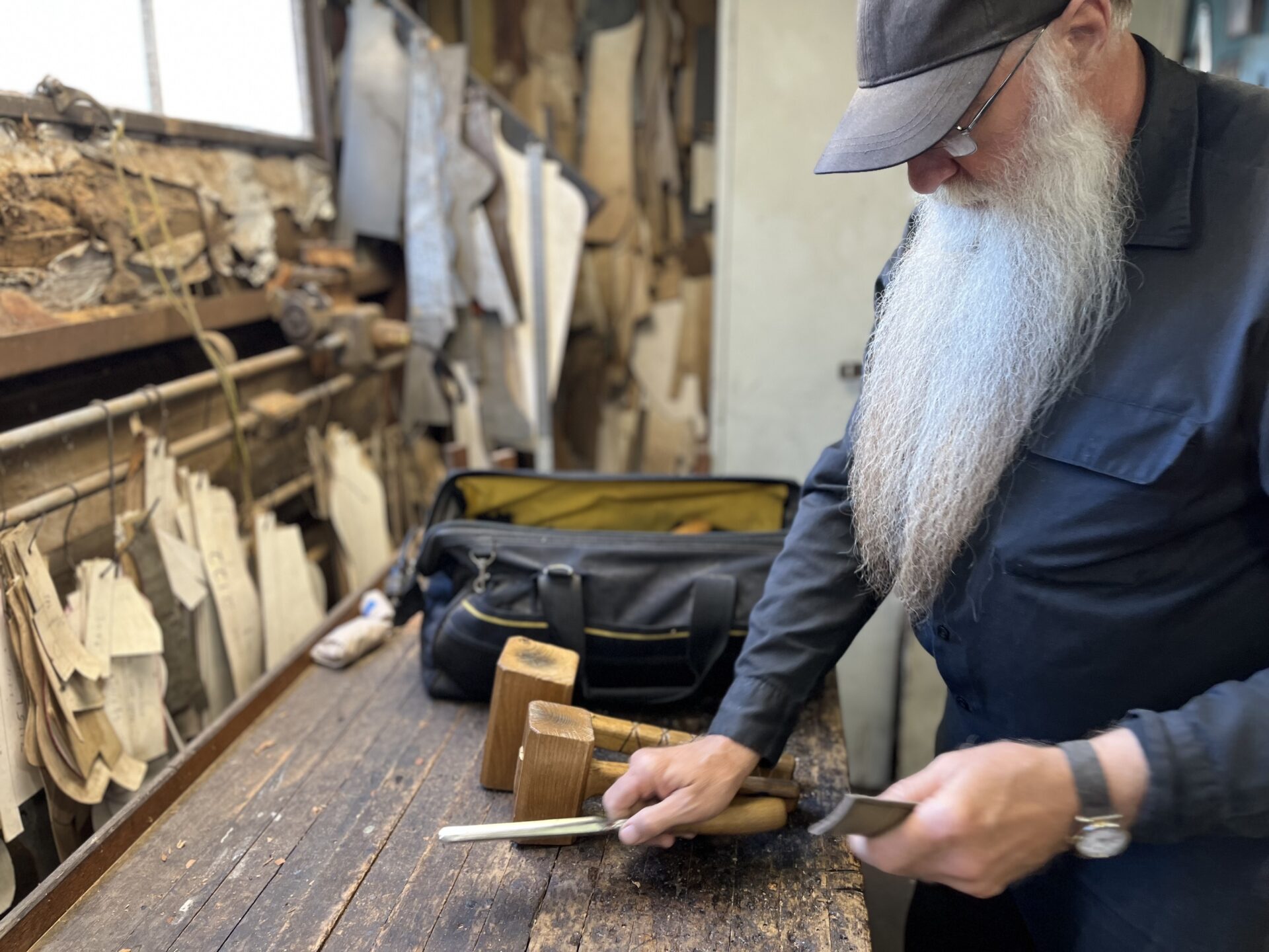 An older factory worker with a long white beard lays out wood carving tools on a brown table.