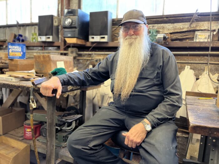 A man with a long white beard and wearing work overalls sits at a workbench.