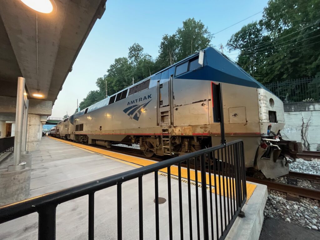 A locomotive pauses at a relatively new concrete station platform in fading daylight on a summer evening.