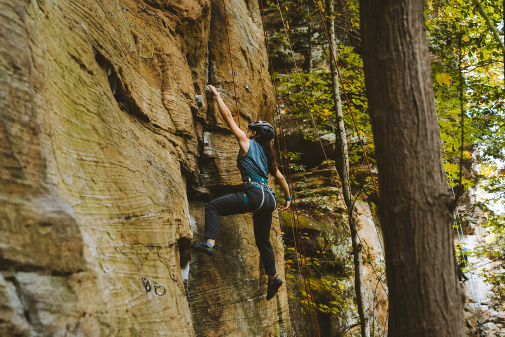 A female rock climber is shown climbing the side of a cliff. She wears a blue shirt and dark pants.
