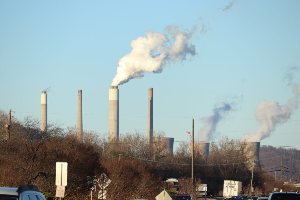 Gray smokestacks and cooling towers rise above a valley on a clear fall day over a bank of leafless trees.