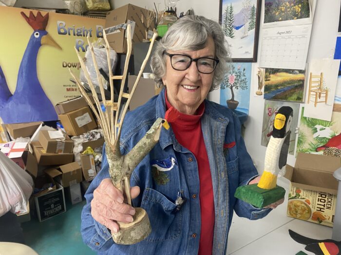 An elderly woman smiles and holds up two wooden art pieces.