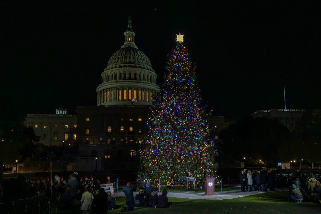 A night image shows a tall evergreen tree wreathed in lights standing in front of the U.S. Capitol, just to the right of the dome. People can be seen gathered at the base of the tree