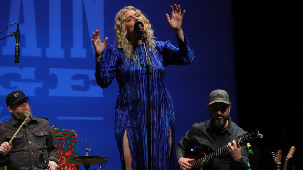 A woman with blond curly hair and a long blue dress sings into a microphone with her hands up and palms facing out.