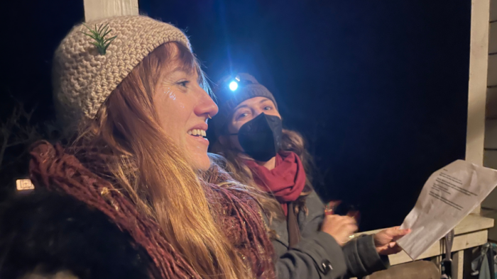 Two carolers sing at night. They look happy and are bundled up in coats, scarves, and hats to keep warm.