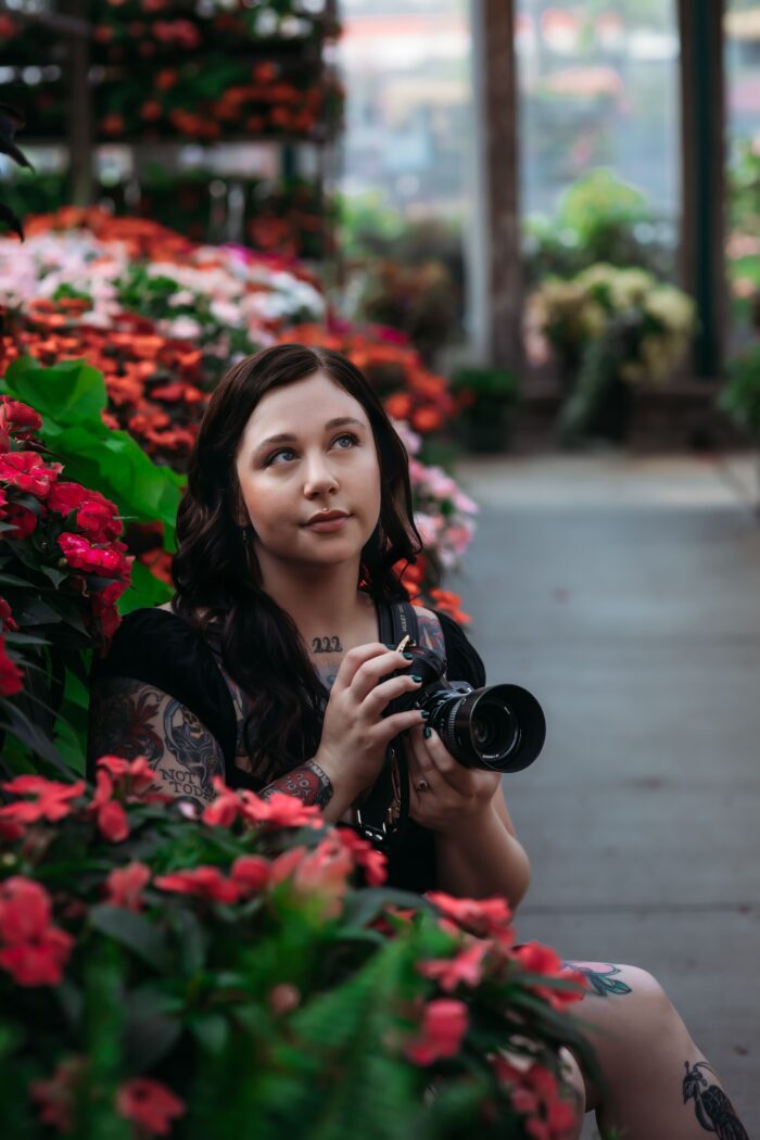 An adult woman with dark hair and tattoos poses for a photo and looks up toward the sky, while holding her camera. A flower bush is seen behind her.